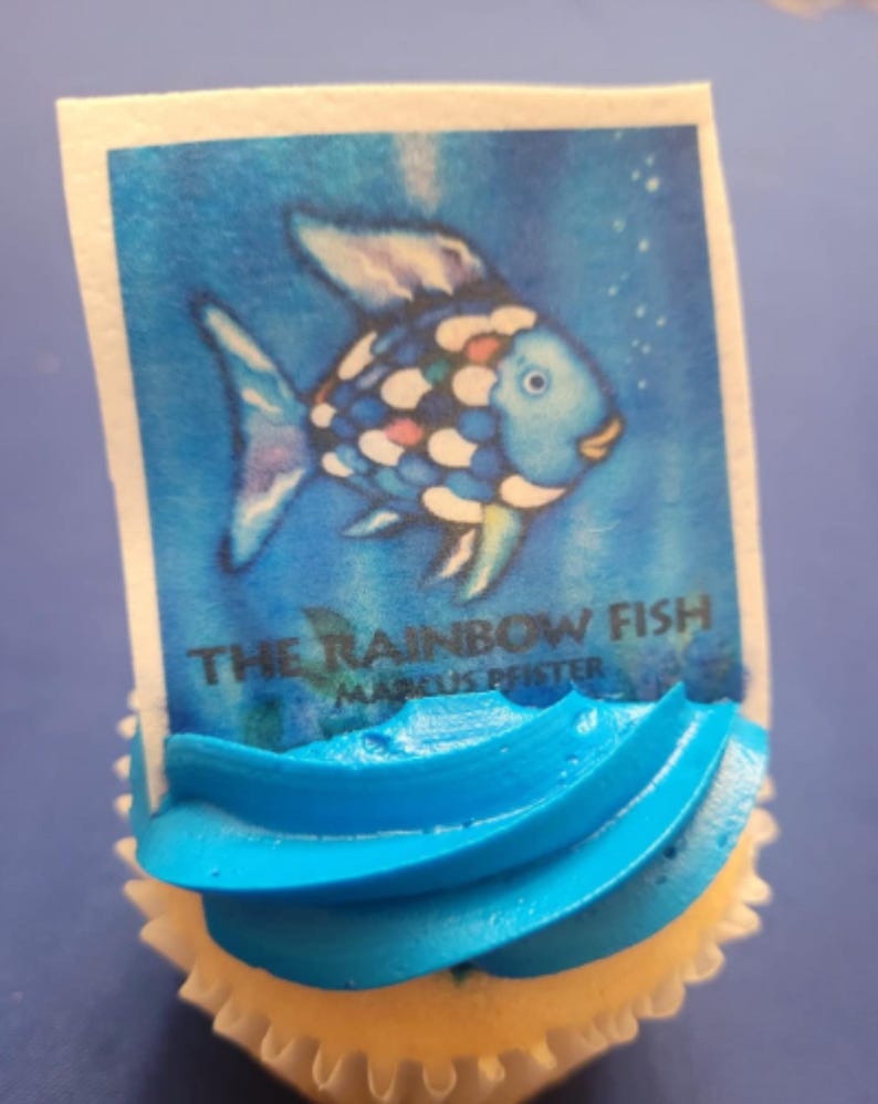 K&ouml;nnte beinhalten: Ein Cupcake mit blauer Glasur und einem rechteckigen essbaren Bild von "The Rainbow Fish" von Marcus Pfister. Das Bild zeigt einen blauen Fisch mit bunten Schuppen vor blauem Hintergrund. Der Cupcake befindet sich in einer wei&szlig;en Papierform.