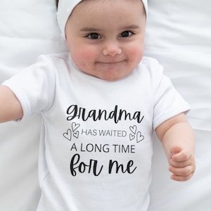 May include: A white baby onesie with black text that reads "Grandma has waited a long time for me" with small hearts around the text.