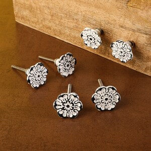 May include: Six white ceramic drawer knobs with black floral designs. The knobs have a distressed finish and are attached to silver screws.