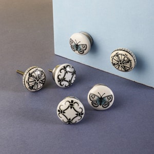 May include: Six white ceramic cabinet knobs with different designs. The designs include a compass, a butterfly, and a geometric pattern. The knobs are all about 1.5 inches in diameter.