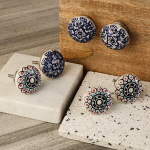 May include: Six ceramic drawer knobs with intricate floral designs. The knobs are white with blue and red accents. The knobs are displayed on a white marble surface.