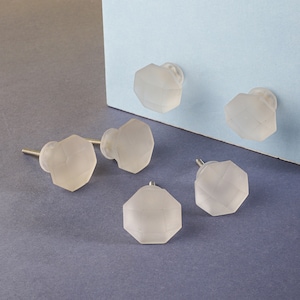 May include: Six frosted glass cabinet knobs in a hexagonal shape. The knobs are a translucent white color and have a silver screw.