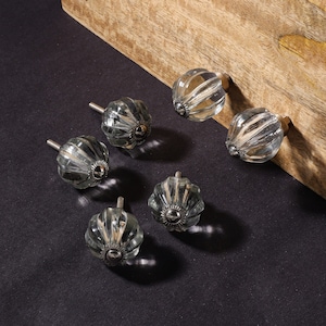 May include: Six clear glass cabinet knobs with silver metal accents. The knobs have a ribbed design and are displayed on a wooden surface.