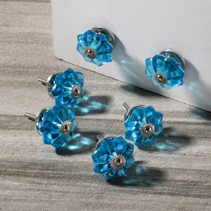 May include: Six blue glass cabinet knobs with silver metal bases. The knobs have a faceted design and are arranged on a light gray surface.