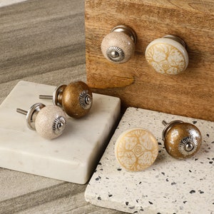 May include: Five ceramic cabinet knobs with a gold and white design. The knobs have a silver metal base and are displayed on a white and brown surface.