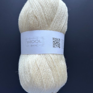 May include: A skein of cream-colored yarn wrapped in a white paper label. The label features the text "WOOL HOUSE" inside a rectangular border, along with a QR code and additional printed information on the right side of the band.