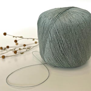 May include: A large ball of pale green yarn with a strand extending outward. The yarn is tightly wound and has a textured appearance. A sprig of dried flowers with small brown buds is to the left of the yarn.