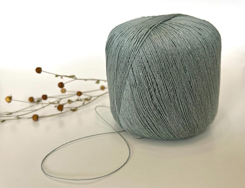 May include: A large ball of pale green yarn with a strand extending outward. The yarn is tightly wound and has a textured appearance. A sprig of dried flowers with small brown buds is to the left of the yarn.