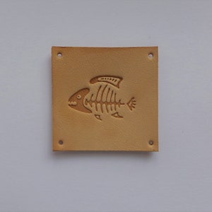 May include: A tan leather patch with four holes and a fish skeleton design embossed in the center.