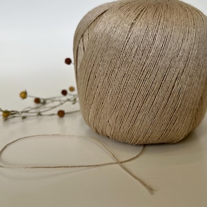 May include: A large ball of beige twine sits on a white surface, with a strand of twine extending from it. Dried flowers with small brown and yellow buds are in the background. The twine is tightly wound.