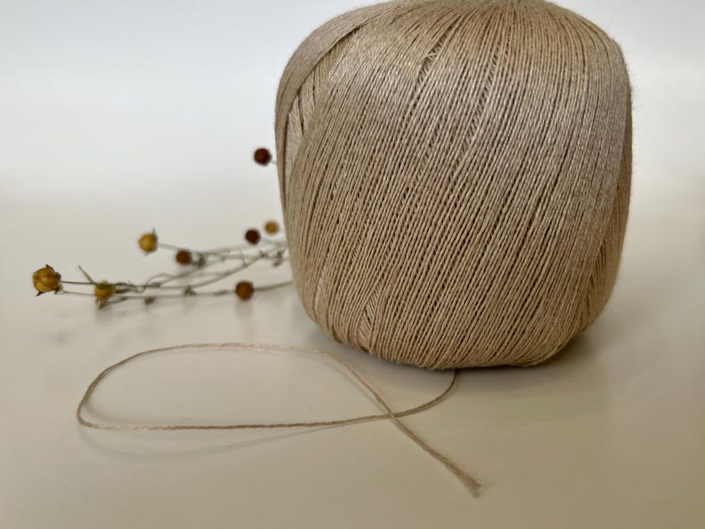 May include: A large ball of beige twine sits on a white surface, with a strand of twine extending from it. Dried flowers with small brown and yellow buds are in the background. The twine is tightly wound.