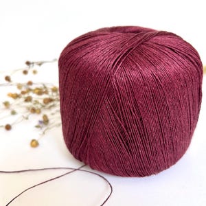 May include: A ball of burgundy yarn sits on a white surface. The yarn is a deep, rich color, with visible strands creating a textured appearance. A few dried flowers are in the background, and a strand of yarn extends from the ball.