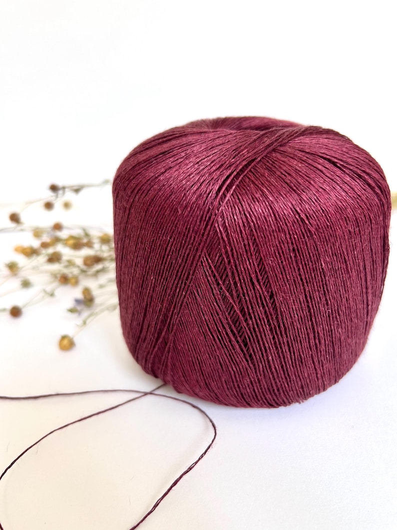 May include: A ball of burgundy yarn sits on a white surface. The yarn is a deep, rich color, with visible strands creating a textured appearance. A few dried flowers are in the background, and a strand of yarn extends from the ball.