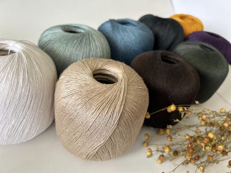May include: A collection of yarn skeins in various colors, including white, beige, blue, black, mustard yellow, and purple. The yarn is arranged in a close-up shot, showcasing the texture and color variations. A few sprigs of dried flowers are in the foreground.