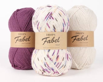 Pode incluir: Três novelos de fio Drops Fabel em várias cores. Um é roxo escuro liso, outro é salpicado com roxo, azul e laranja sobre um fundo creme, e o terceiro é creme liso. Cada novelo tem uma etiqueta de papel castanho.