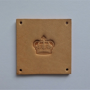 May include: A square, tan leather patch with a detailed embossed crown design in the center. The patch has four evenly spaced holes, one in each corner. The leather has a natural, textured appearance.