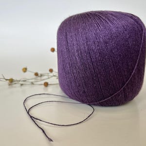 May include: A large ball of deep purple yarn sits on a white surface. A strand of the yarn extends from the ball. Dried, tan-colored sprigs are in the background. The yarn appears to be made of a soft, natural fiber.