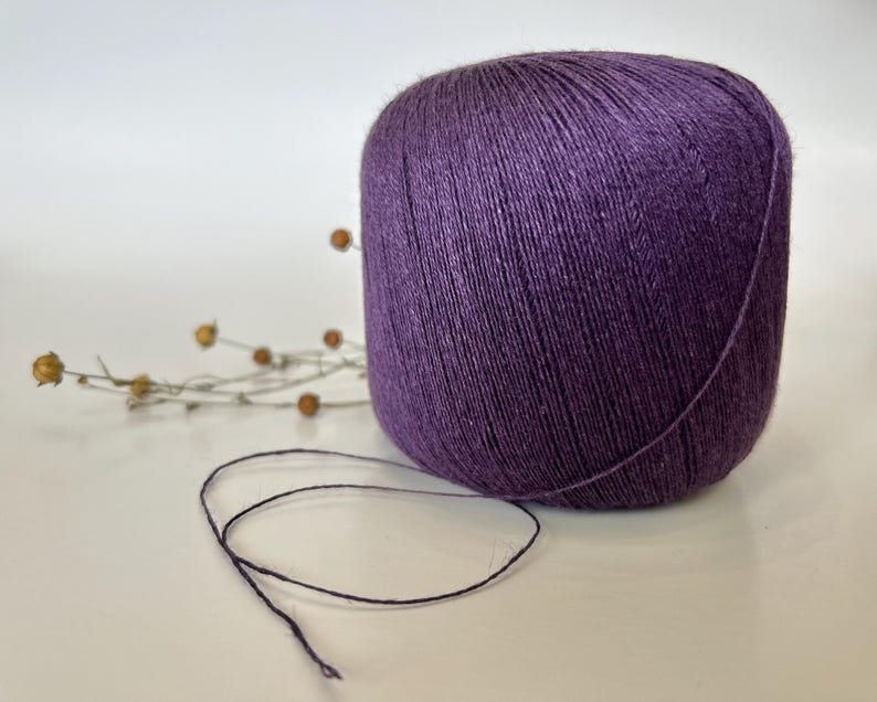May include: A large ball of deep purple yarn sits on a white surface. A strand of the yarn extends from the ball. Dried, tan-colored sprigs are in the background. The yarn appears to be made of a soft, natural fiber.