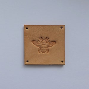 May include: A light brown leather patch with a bee design embossed in the center. The patch has four holes around the edges.