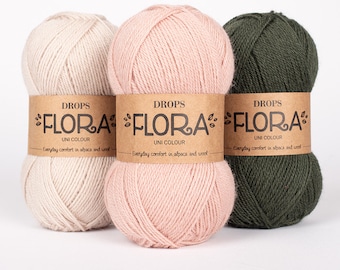 Pode incluir: Três novelos de fio nas cores bege claro, rosa suave e verde floresta escuro. Cada novelo apresenta um rótulo de papel castanho com o texto DROPS FLORA UNI COLOUR e a frase Everyday comfort in alpaca and wool impressa em tinta preta.