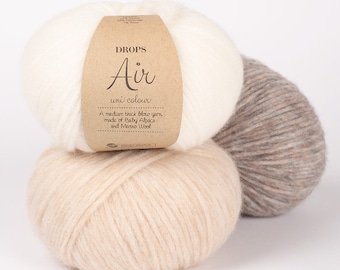 May include: Three balls of Drops Air uni colour yarn in cream, beige, and gray. The cream-colored ball has a brown label with the brand name and product details. The yarn is described as a medium thick blow yarn made of Baby Alpaca and Merino Wool.