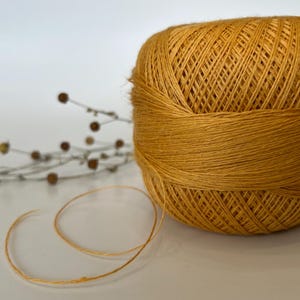 May include: A golden-yellow ball of yarn with a strand unwound, set against a white background. The yarn is tightly wound, showing the texture of the fibers. A sprig of dried flowers is in the background.