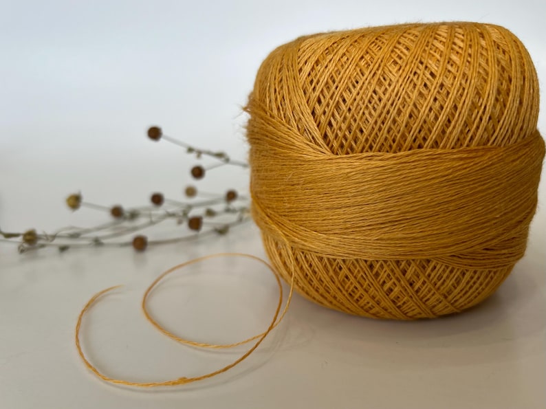 May include: A golden-yellow ball of yarn with a strand unwound, set against a white background. The yarn is tightly wound, showing the texture of the fibers. A sprig of dried flowers is in the background.