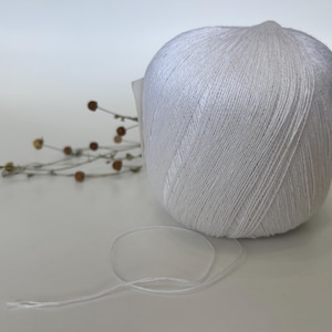 May include: A large ball of white yarn with a loose strand extending from it. The yarn is tightly wound, and the texture of the individual threads is visible. A few dried stems with small brown buds are in the background.