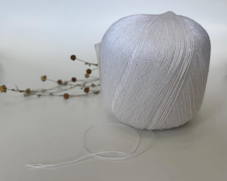 May include: A large ball of white yarn with a loose strand extending from it. The yarn is tightly wound, and the texture of the individual threads is visible. A few dried stems with small brown buds are in the background.