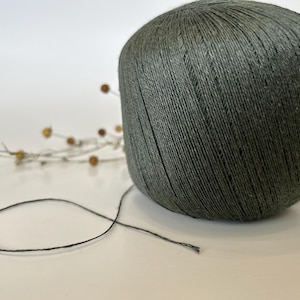 May include: A large ball of dark gray yarn with a strand extending outward. The yarn is tightly wound, and the texture of the individual threads is visible. A sprig of dried flowers is in the background.