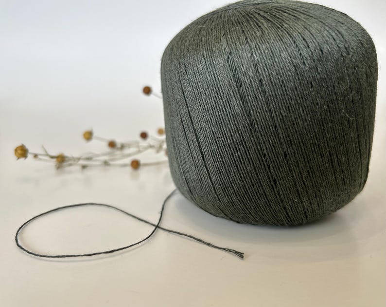 May include: A large ball of dark gray yarn with a strand extending outward. The yarn is tightly wound, and the texture of the individual threads is visible. A sprig of dried flowers is in the background.