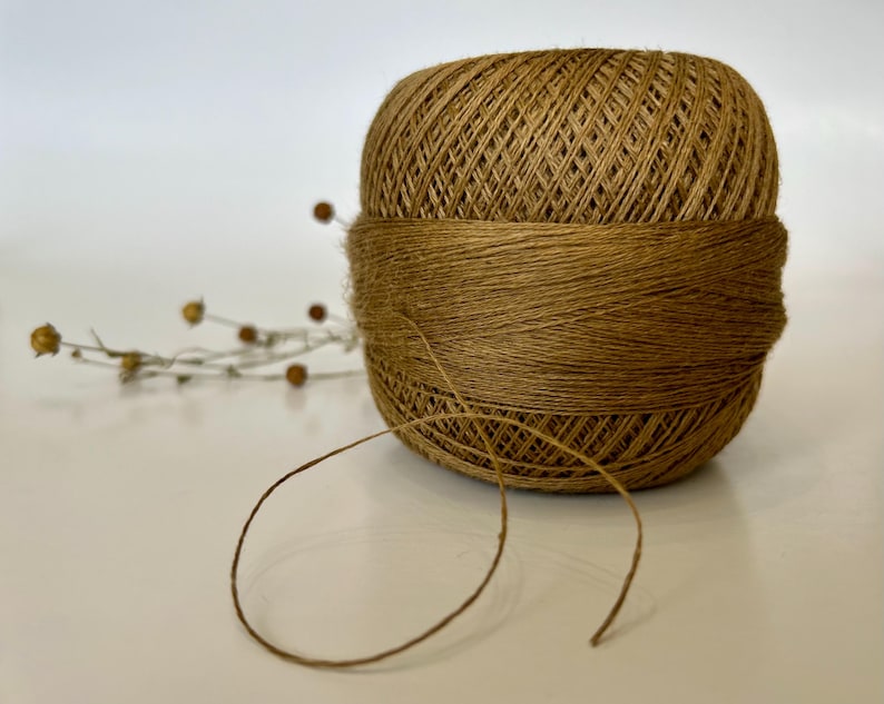 May include: A ball of brown twine, tightly wound, with a band of the same material wrapped around its center. A strand of twine extends from the ball. Dried sprigs with small, round seed pods are placed to the left of the twine ball.
