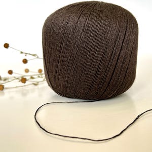 May include: A large ball of dark brown yarn with a strand extending from it. The yarn is tightly wound, and the texture is visible. A few sprigs of dried flowers are in the background, suggesting a craft or hobby theme.
