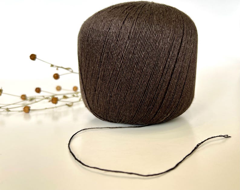 May include: A large ball of dark brown yarn with a strand extending from it. The yarn is tightly wound, and the texture is visible. A few sprigs of dried flowers are in the background, suggesting a craft or hobby theme.