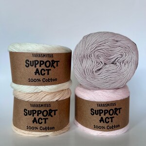 May include: Four spools of yarn in shades of white, peach, pink, and lavender. Each spool has a brown paper label with the text "YARNSMITHS SUPPORT ACT 100% Cotton". The yarn is neatly wound, ready for crafting.