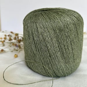 May include: A large spool of olive green yarn. The yarn is tightly wound, with visible individual strands. A length of the yarn extends from the spool, suggesting it's ready for use in knitting or crochet projects. The background is a soft, neutral color.