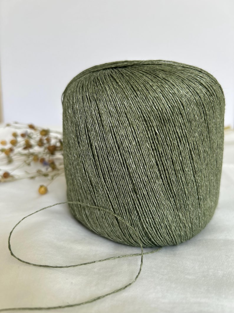 May include: A large spool of olive green yarn. The yarn is tightly wound, with visible individual strands. A length of the yarn extends from the spool, suggesting it's ready for use in knitting or crochet projects. The background is a soft, neutral color.