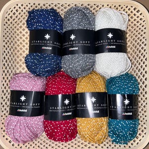 May include: Assortment of eight skeins of sparkly yarn in various colors, including pink, red, gold, teal, white, gray, and blue. Each skein has a label that reads "Starlight Soft" with a star logo and the brand name "Hobbi".