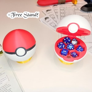 May include: A red and white Pokeball-shaped dice tower with a blue and white dice set inside. The dice tower is open and the dice are visible. The dice tower is on a white surface. The text "*Free Stand!" is written in black on a white background.
