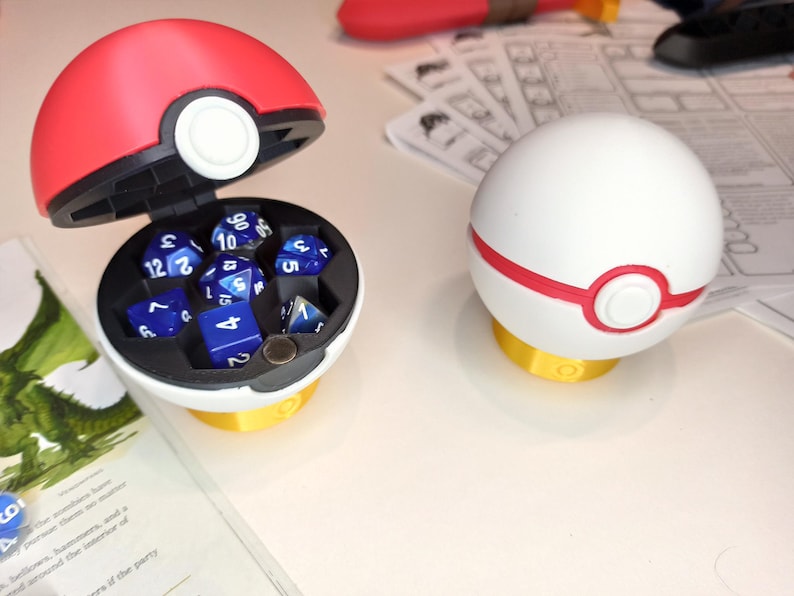 May include: A red and white Pokeball-shaped dice container with a lid that opens to reveal a compartment for dice. The container is filled with blue and white dice with numbers on them.