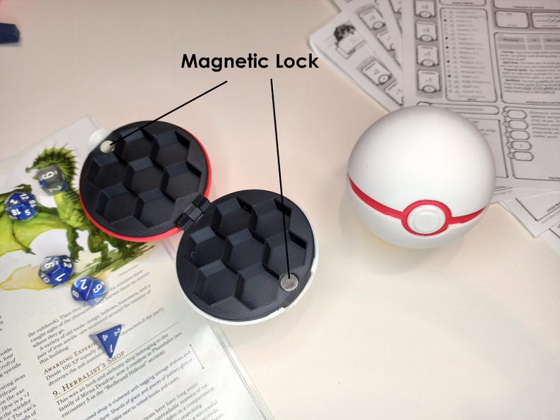 May include: A red and black dice rolling box with a magnetic lock. The box is open and has a black honeycomb interior. A white sphere with a red band and a red circle in the center is next to the box.