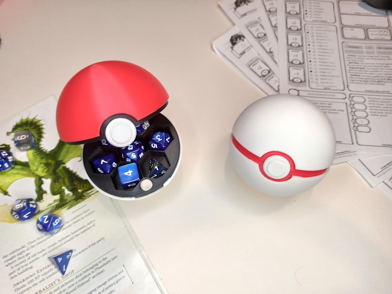 May include: Two Pokeball-shaped dice containers, one red and one white, with a red ring around the center. The red Pokeball is open and contains blue dice with white numbers.