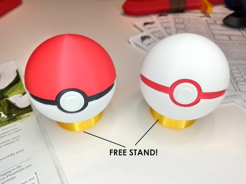 May include: Two Pokeball-style spheres, one red and one white, with a black ring and a red ring, respectively. Both spheres have a white circle in the center. The spheres are on a gold stand. The text "FREE STAND!" is written below the spheres.