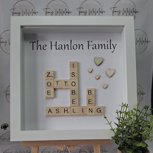 May include: A white frame with a white mount and a white background. The frame contains a scrabble tile word art piece that spells out "The Hanlon Family" using the letters from the scrabble tiles. There are also small wooden hearts scattered around the word art.