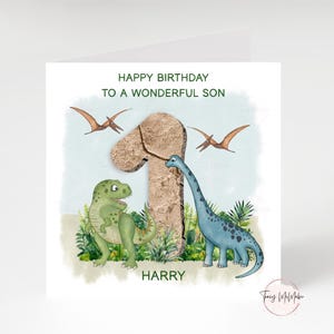 Peut inclure: Carte d'anniversaire avec un dinosaure vert et orange, un dinosaure bleu et deux ptérosaures volants. La carte porte l'inscription "JOYEUX ANNIVERSAIRE À UN FILS MERVEILLEUX" et le nom "HARRY". Le chiffre "1" est en arrière-plan.