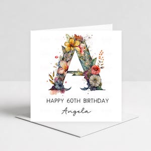 Peut inclure: Carte de vœux blanche avec un motif floral en forme de lettre "A". La lettre est ornée de fleurs et de feuillages colorés. La carte indique "JOYEUX 60E ANNIVERSAIRE Angela" en texte noir.