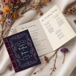 May include: A navy blue wedding order of service booklet with a burgundy spine and gold floral accents. The front cover features the text "Order of Service for the Wedding of Willow & James 27.10.2022". The back cover lists the wedding party members and their roles.