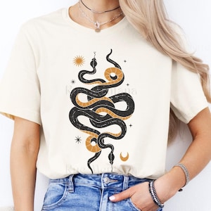 Op de afbeelding: Een wit T-shirt met een zwart en gouden slangontwerp. De slang is opgerold en heeft een halve maan en sterren op de achtergrond.