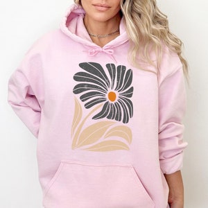 K&ouml;nnte beinhalten: Ein rosa Hoodie mit einem schwarzen und beigefarbenen Blumendesign. Die Blume hat eine gro&szlig;e, dunkle Mitte mit einem kleinen orangefarbenen Kreis in der Mitte.