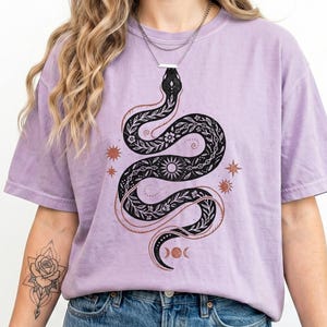 Op de afbeelding: Een lila T-shirt met een zwart slangenontwerp, met bloemen en hemelse elementen. De slang is opgerold, met een zon en maanfasen in het ontwerp. Het shirt heeft een relaxte pasvorm en korte mouwen. Het woord "Orchid" is zichtbaar.